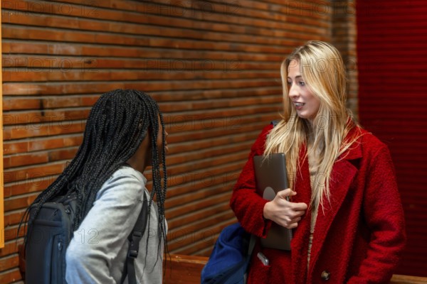Two female students chatting during break time in university corridor, holding laptop and backpack