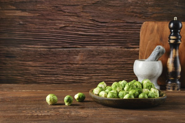 A wooden bowl filled with fresh Brussels sprouts sits on a rustic wooden table. Nearby, a mortar,