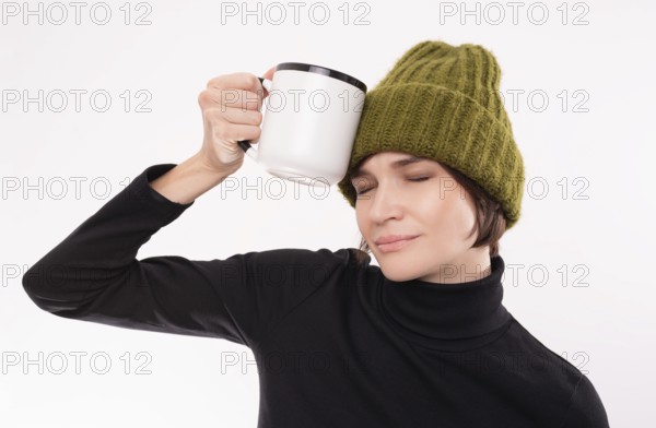 Funny girl in a green knitted hat put a white mug to her temple. The concept of a hangover after