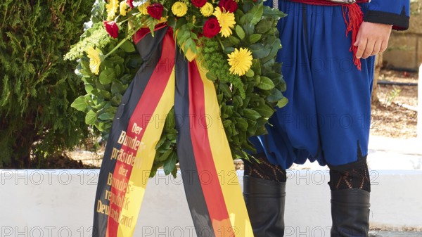 Wreath of flowers in the colours of Germany with a man dressed in traditional Cretan costume, visit