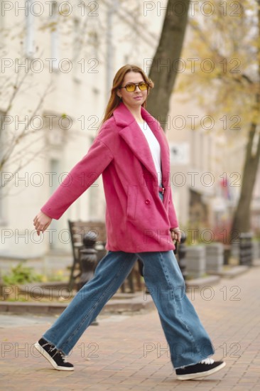 A person walks confidently along a tree-lined street, wearing a pink coat, sunglasses, and