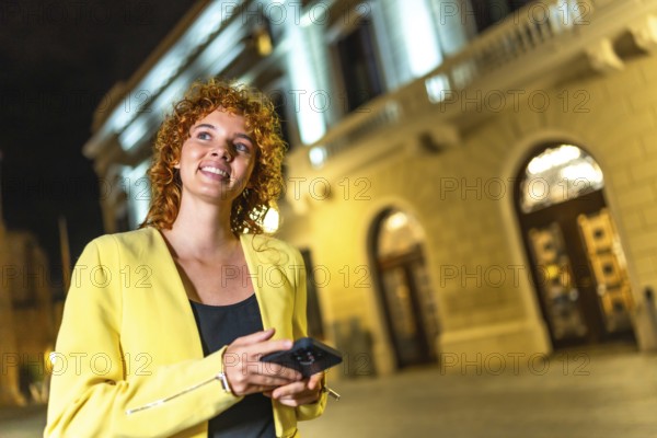 Happy woman in casual colorful clothes and curly hair looking at city views using phone at night