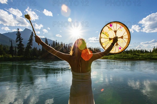 A close up and rear view of a shaman woman holding a sacred powwow drum towards sun and mountains