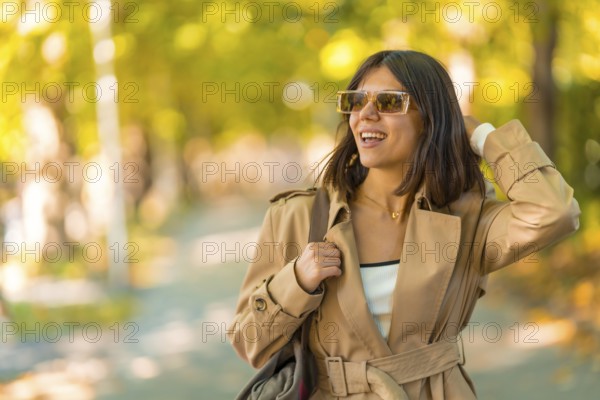 A woman wearing a tan coat and sunglasses is walking down a path. She is smiling and she is