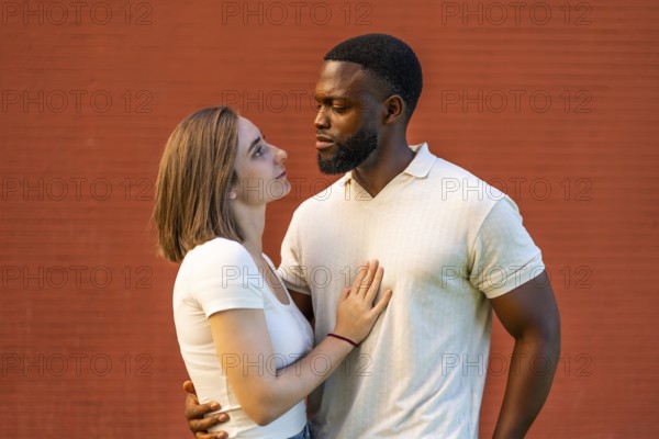 Mixed-raced couple standing embracing and looking at each other against red brick wall