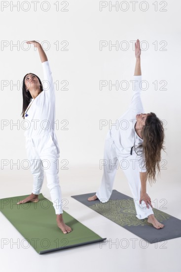 Two women dressed in white doing yoga over white background. Utthita trikonasana yoga pose. Studio