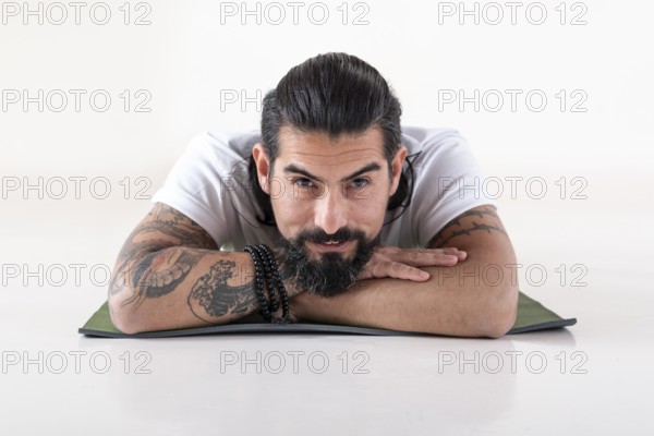 Portrait of a man dressed in white yoga clothes lying on a mat while looking at camera over white