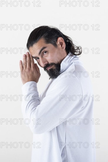 A man dressed in white clasping hands while looking at camera over white background. Tadasana yoga