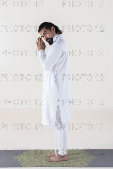 A man dressed in white on a mat, clasping hands while looking at camera over white background.