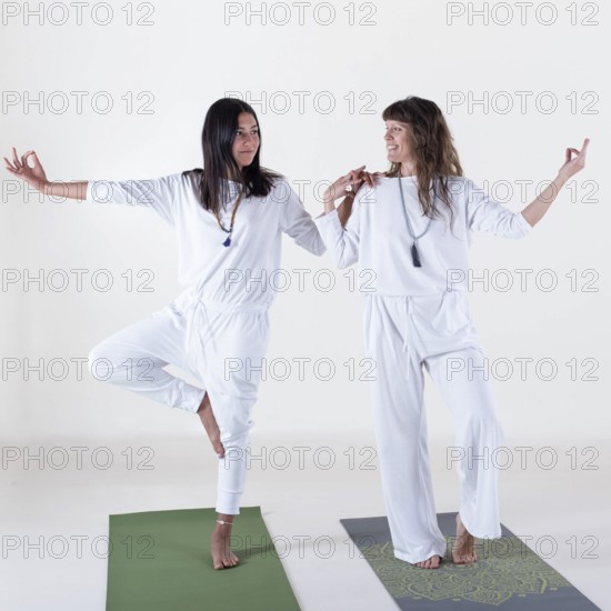 Two women dressed in white doing yoga over white background. One is teaching the other. Studio shot