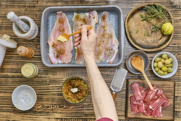 Fresh fish fillets are being seasoned in a rustic kitchen. Herbs, spices, and olive oil are neatly