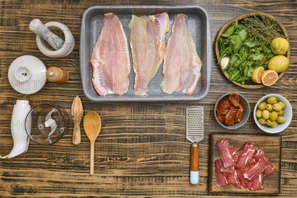 Various fresh ingredients are arranged on a wooden tabletop, including fish fillets, herbs, olives,