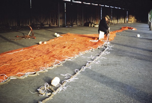 Man sitting in a storage shed mending a fishing net, unspecified location, Great Britain 1975