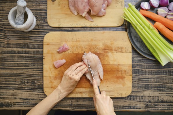 Hands skillfully cut raw chicken fillets on a wooden cutting board surrounded by fresh vegetables