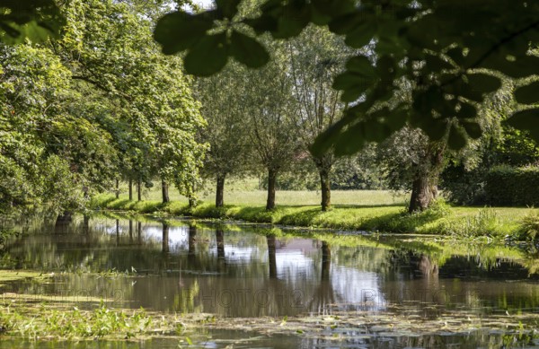 A calm river flows through a green landscape, lined with trees that are reflected in the water,