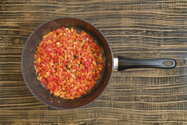 Brightly colored bell peppers are chopped and sauteing in a frying pan on a rustic wooden surface.