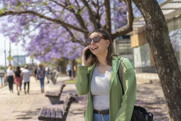 A cheerful young woman with sunglasses and a green jacket talks on her phone during a sunny day on