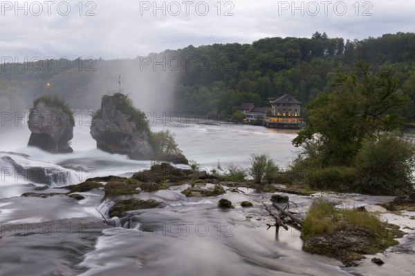 Impressive waterfall with two rocks, surrounded by forest and a building in the background in