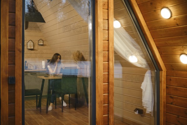 View from outside at a dreamy woman sitting at a table in log cabin in the evening