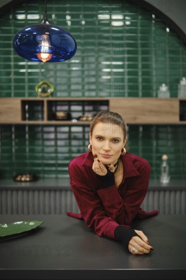 A woman is leaning on a kitchen counter, wearing a red outfit with long sleeves. The background