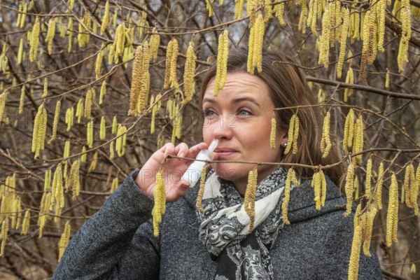 Young Caucasian woman with allergy problem stands by flowering hazel bush and sprays her nose in