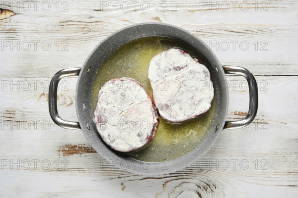 Overhead view of two floured steaks are sizzling in a pan with oil. The pan is on a white wooden