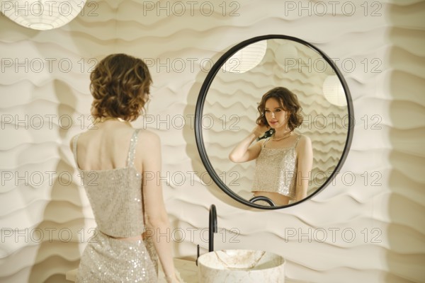 A woman dressed in a shimmering top stands in front of a round mirror, applying makeup. The stylish