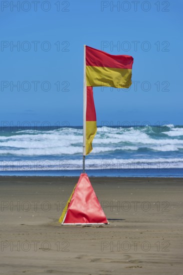 A red and yellow coastguard banner on the beach against a backdrop of waves under a clear blue sky,