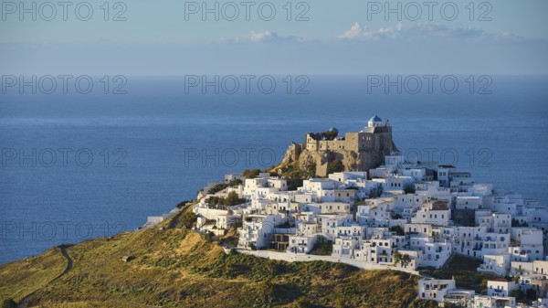 Panorama with white buildings on a hill, dense waves surrounding an old fortress by the sea,