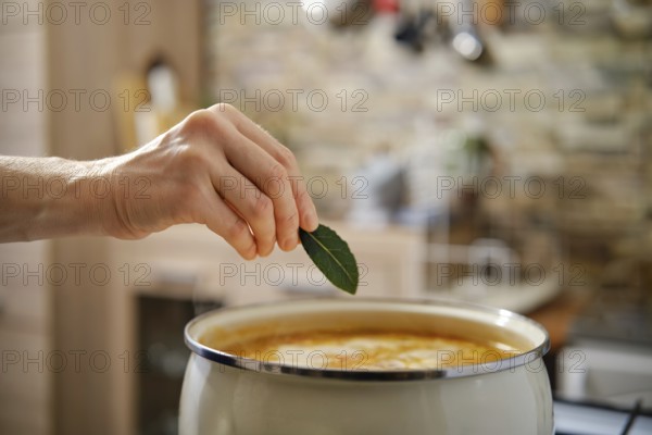 A hand reaches to place a fresh bay leaf into a pot of flavorful soup. The warm kitchen setting