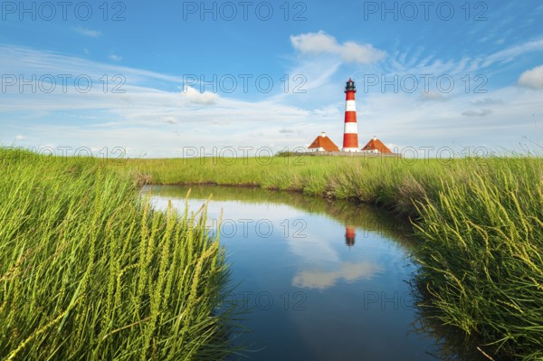 The Westerheversand lighthouse under a blue sky with white clouds reflected in a tideway,