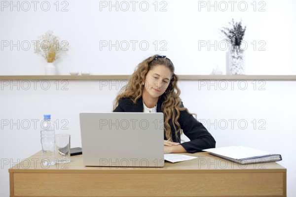 A young woman sits at a contemporary wooden desk, engaged in work on her laptop. Papers and a water