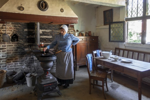 Old woman cooking on antique Flemish cast-iron coal stove, Leuvense stoof in 19th century house,