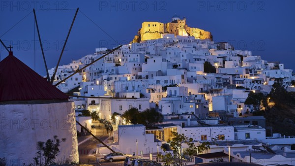 A windmill and illuminated streets lead to an illuminated fortress above white buildings, Querini