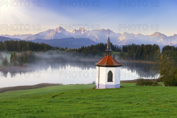 Small chapel on the lake in front of an impressive mountain backdrop at sunrise, surrounded by