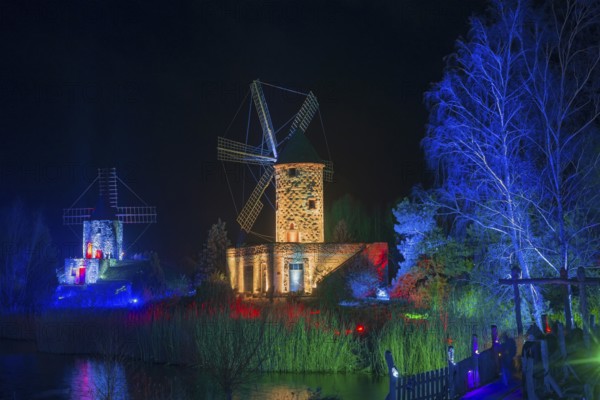 Two windmills in a nocturnal scene, illuminated with colourful lights and surrounded by water,