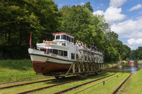 A boat being transported overland on a railway wagon, surrounded by forest and blue sky, ship