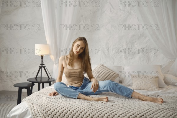 A young woman sits comfortably on a spacious bed in a serene, light-colored room. Her relaxed