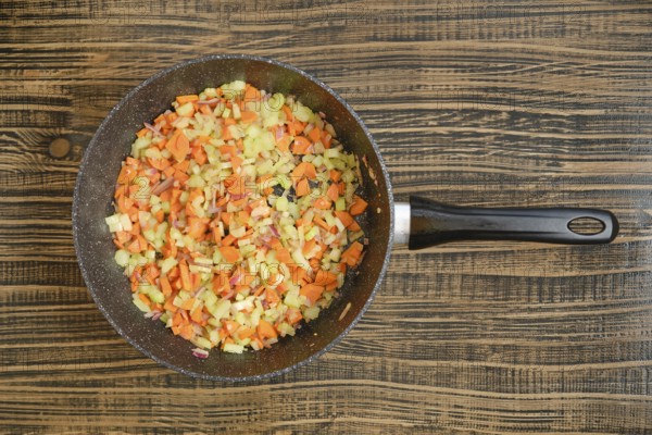 Colorful chopped carrots, onions, and celery are cooking in a black skillet on a wooden table. The
