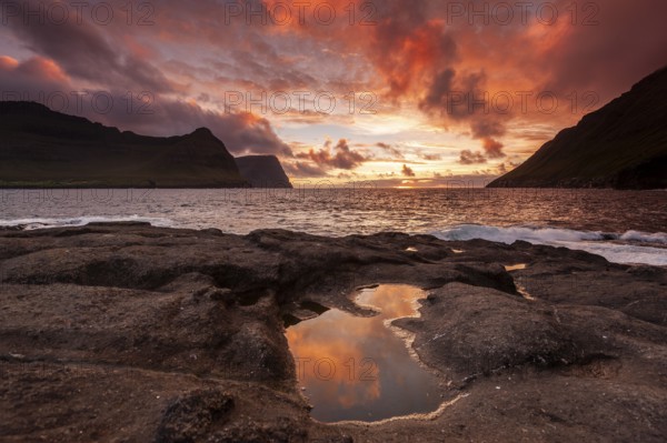 Sunset over the sea between steep mountain slopes, reflection in calm pool on rocks, Vidareidi,