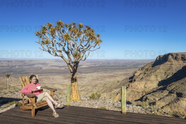 A woman sits with a cup of tea in a chair on the terrace of the Fish River Lodge, behind her the
