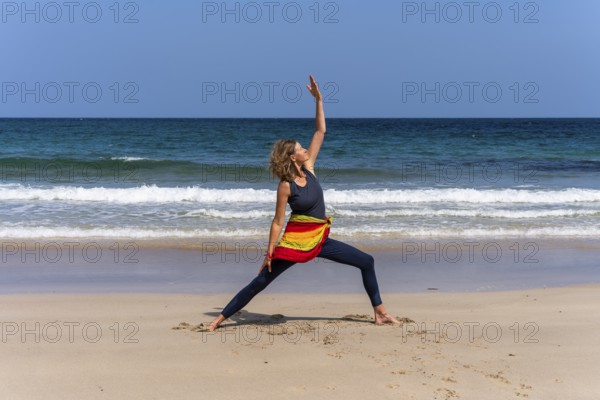 A woman doing yoga on a sandy beach by the sea. Shanti Virabhadrasana or Peaceful Warrior. Treen