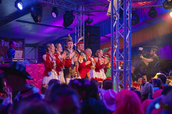 A group on stage at a carnival event, surrounded by a celebrating crowd, Fasching, Schlagerkuchen