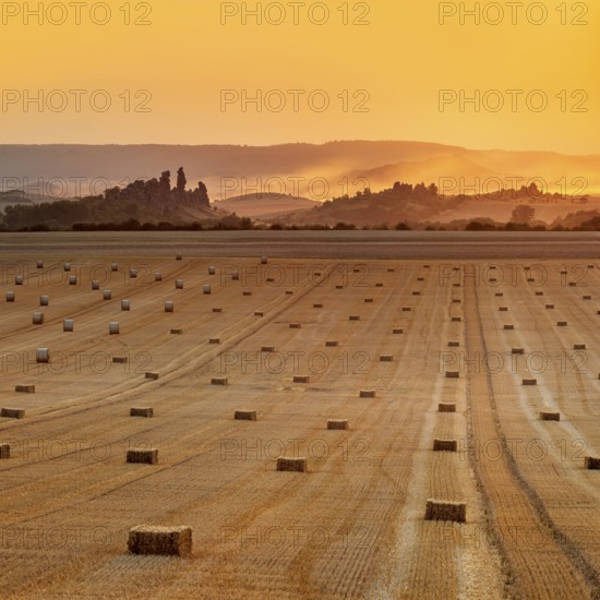View over stubble field with straw bales to the Devil's Wall at sunset, fog rising, Weddersleben,