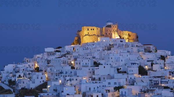 An illuminated fortress towers above white-washed buildings under a deep blue night sky, Querini