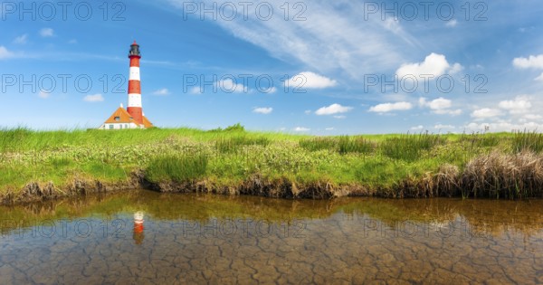 The Westerheversand lighthouse under a blue sky with white clouds reflected in a tideway,