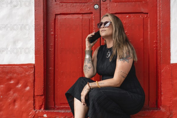 A stylish tattooed woman, dressed in a black dress and sunglasses, holds her smartphone while