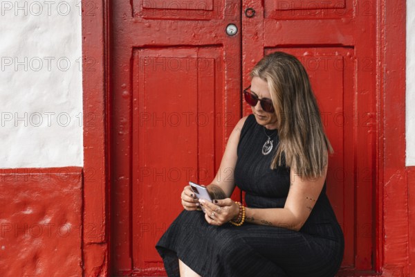 A tattooed woman in sunglasses and black dress is sitting in front of a vibrant red door,