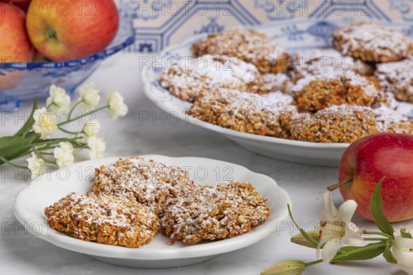 Sugar-dusted oat biscuits on a plate surrounded by apples and flowers in front of patterned tiles