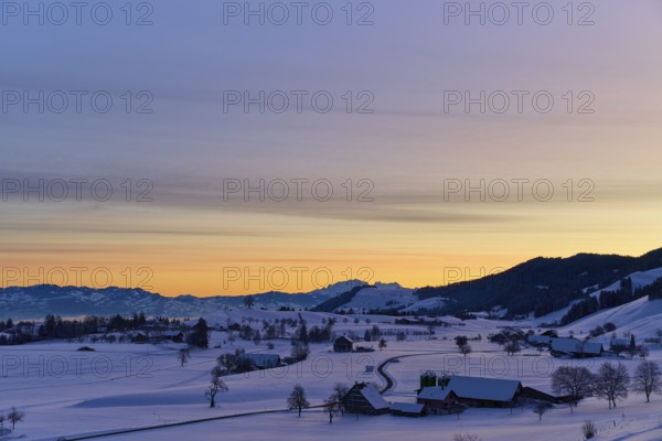 Snowy landscape at sunrise with mountains and farms, bathed in a warm orange-blue light, Menzingen,